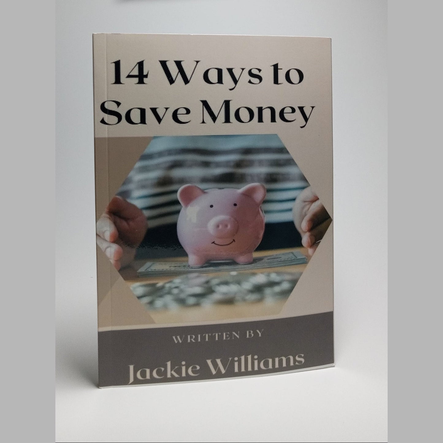 A book cover titled '14 Ways to Save Money' by Jackie Williams, featuring a pink piggy bank and hands counting coins on the front, shown standing upright against a light gray background.